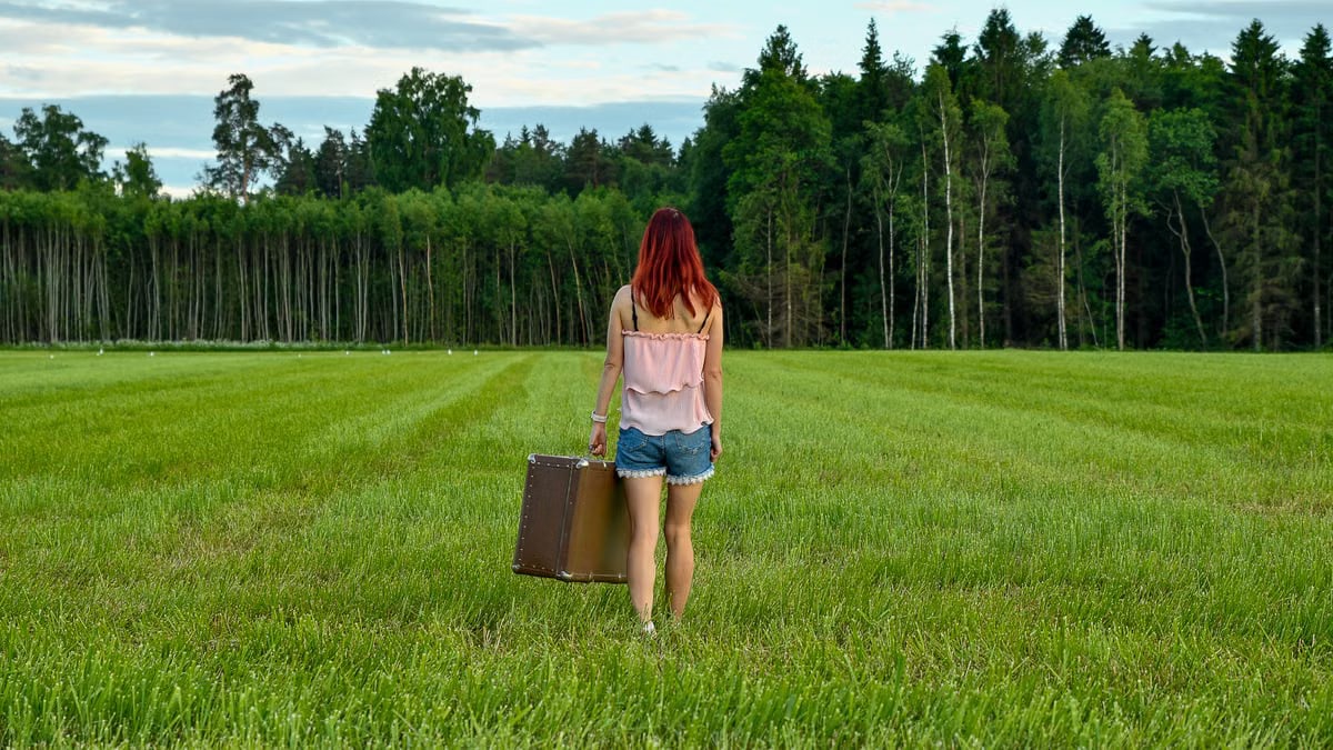 A woman in denim shorts and pink top walks through a meadow carrying a vintage suitcase, symbolizing new beginnings.